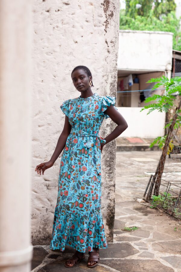 Dress made from hand block printed cotton in soft hues, featuring pleated details, a round neckline, and off-shoulder ruffles. The slim-fitted bodice hugs curves, flowing into a floor-length skirt. Includes a detachable belt and inseam pockets.