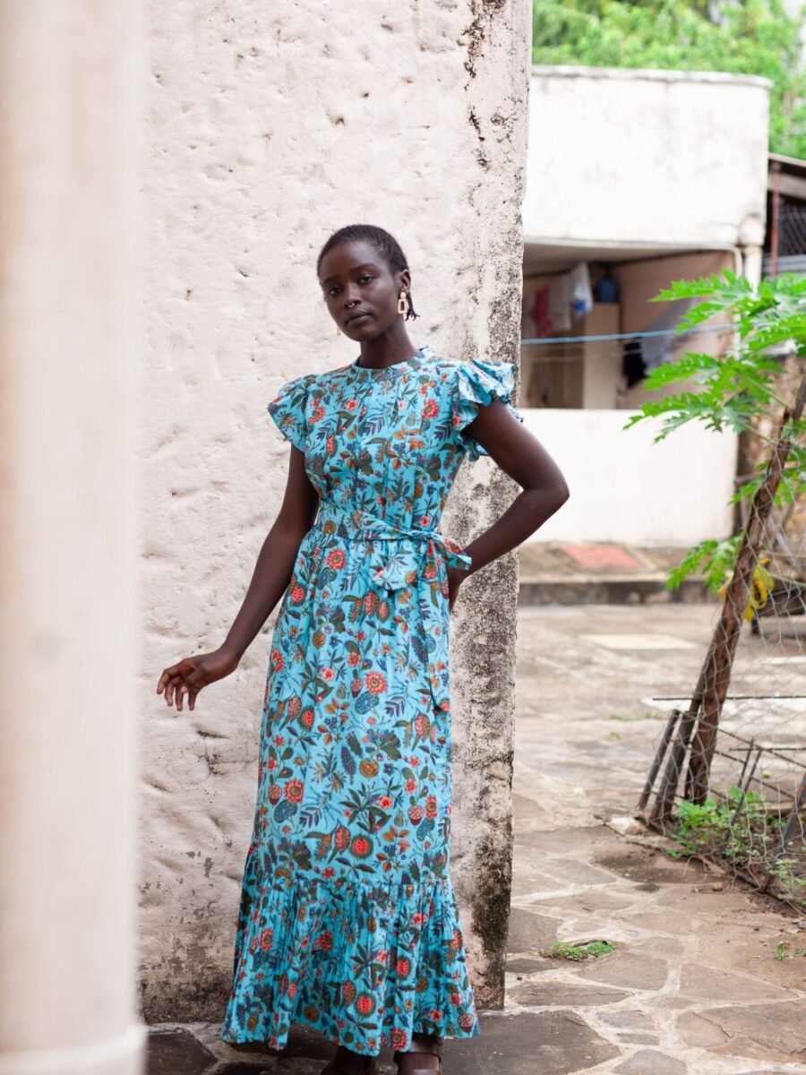 Dress made from hand block printed cotton in soft hues, featuring pleated details, a round neckline, and off-shoulder ruffles. The slim-fitted bodice hugs curves, flowing into a floor-length skirt. Includes a detachable belt and inseam pockets.